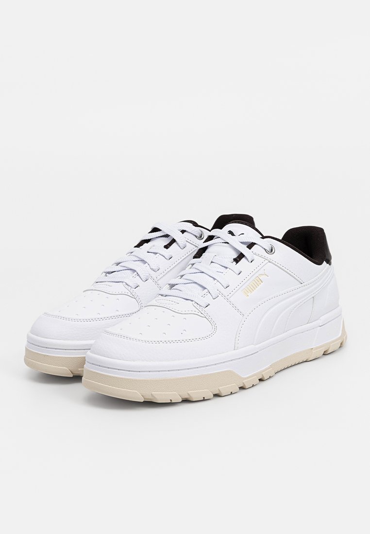 Para białych, niskich sneakersów Puma z beżowymi podeszwami, perforowaną strefą na palce, białymi sznurowadłami i czarną wewnętrzną wyściółką na białym tle.