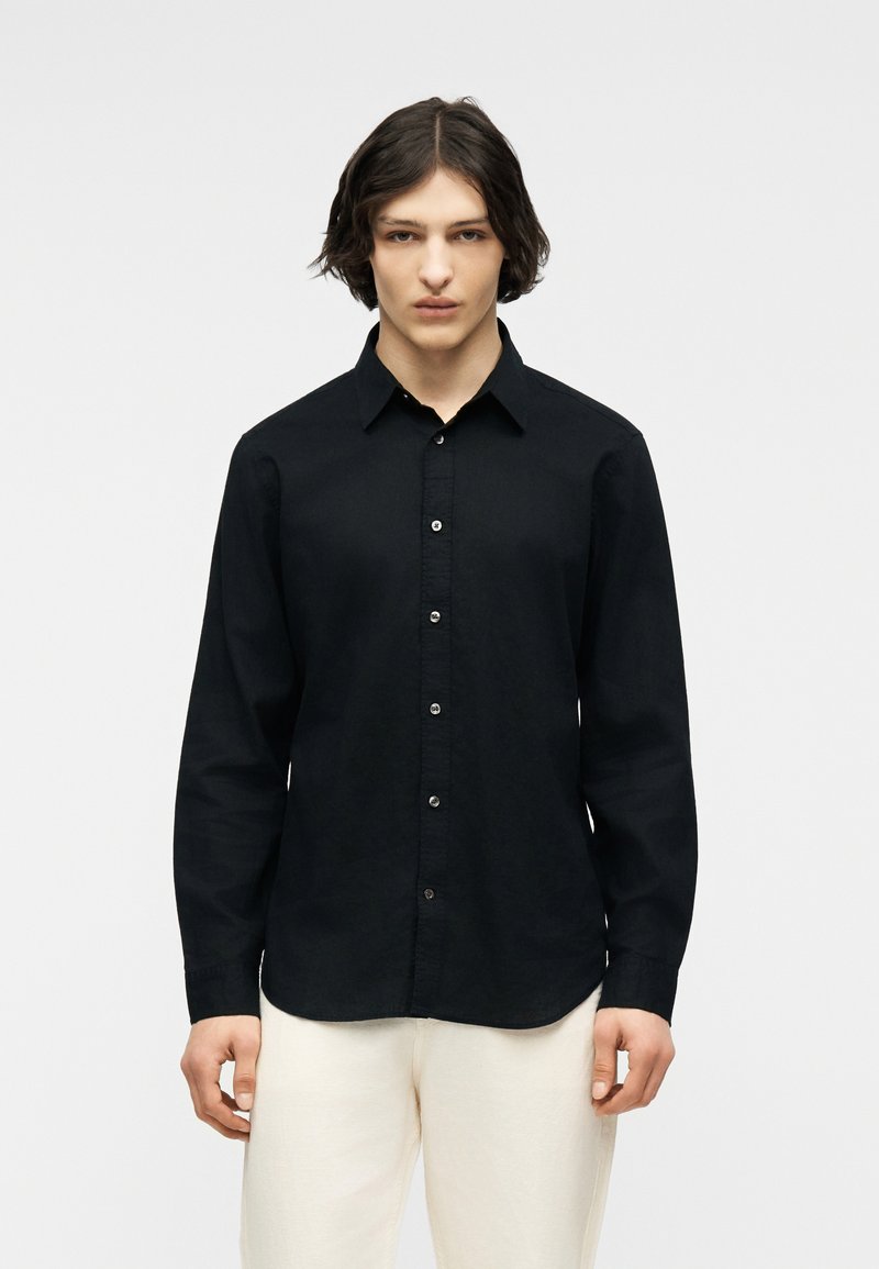 Jeune personne aux cheveux courts et foncés portant une chemise noire à manches longues boutonnée et un pantalon de couleur claire, debout devant un fond uni.