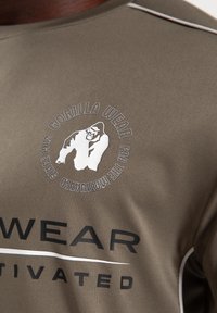 Olijfgroen kortebloesemshirt van gladde stof. Voorzien van een wit logo met een gorillagrafiek en zwarte tekst, geaccentueerd met grijze stiksels.