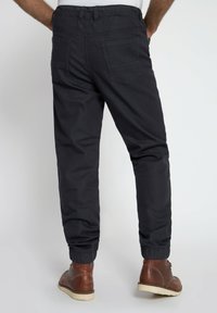 Pantaloni jogger neri con vita elastica, fondo a costina, due tasche posteriori e dettagli di cucitura minimi. Indossati con stivali marroni.