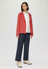Koraalkleurige woljacket met een notch-kraag en twee knopen, gecombineerd met een witte blouse, donkere denim wijde broek en crèmekleurige instappers.