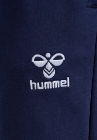 Marineblaue Hose aus weichem Stoff mit einem weißen gestickten Logo in stilisiertem Design, platziert unten links.