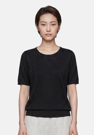 Jeune femme aux cheveux courts et foncés portant un t-shirt noir uni à manches courtes et un pantalon beige clair, debout devant un fond blanc.
