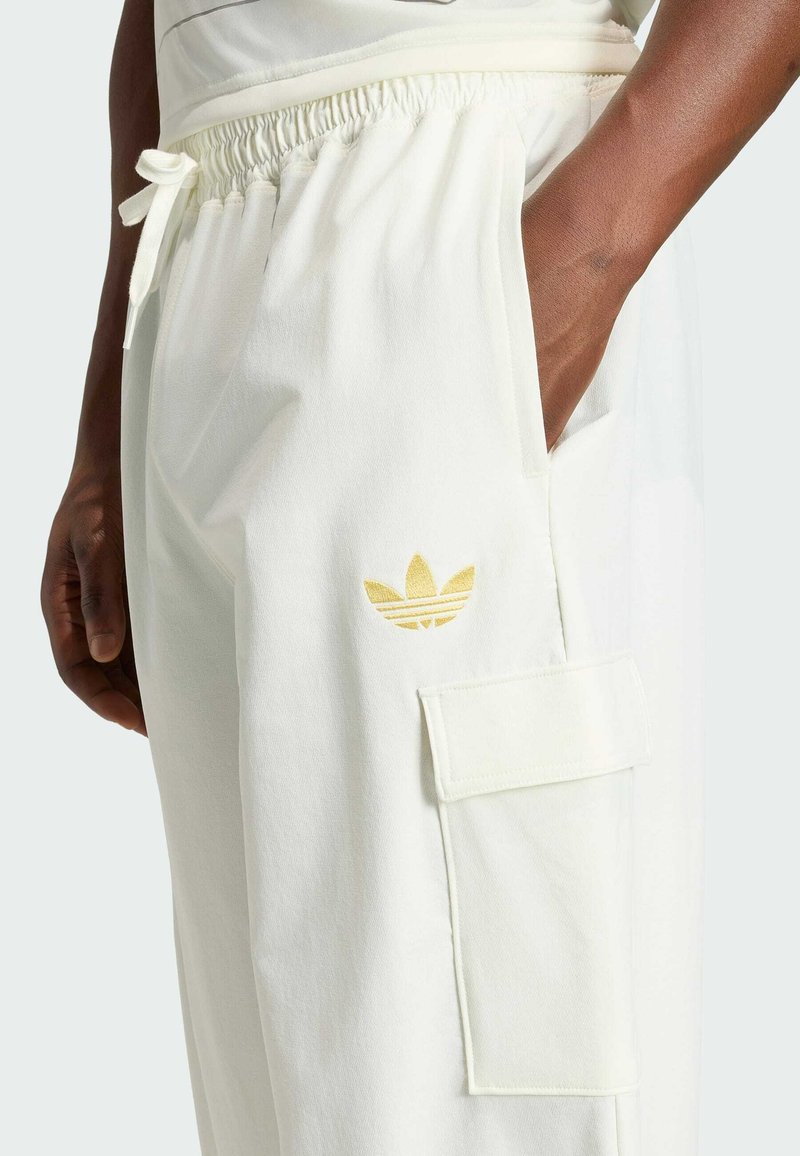 Person mit dunkler Hautfarbe, die weiße Jogginghosen mit elastischem Bund, Seiten- und Cargotaschen sowie einem kleinen goldenen Adidas-Logo in der Nähe des Oberschenkels trägt.