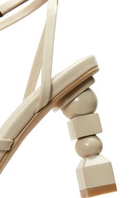 Beige lakleren sandaal met een brede enkelband, voorzien van een sculpturaal blokhak dat bestaat uit twee bollen en een vierkante basis.