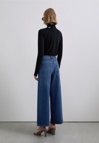 Top nero a maniche lunghe con collo alto, abbinato a jeans azzurri a gamba larga e sandali argento con tacco alto. Vista posteriore con un sottile dettaglio a ritaglio.