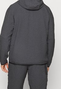 Man klädd i en mörkgrå hooded jacka med textur och matchande byxor, sedd bakifrån mot en enkel ljus bakgrund.