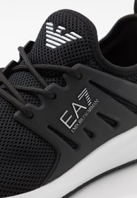 Černá sportovní bota s prodyšným síťovaným svrškem, s texturovaným syntetickým překrytím, zavazováním na tkaničky a bílým logem na boku.