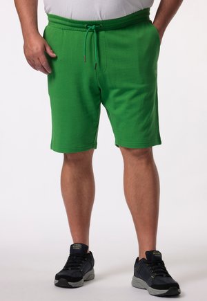 Homme portant un short vert avec un cordon, une chemise blanche et des chaussures de sport noires, debout devant un fond blanc uni.
