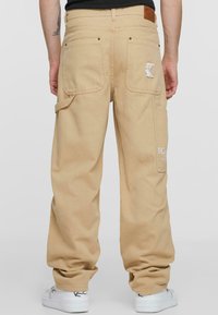Beige cargo-housut, joissa on kaksi takataskua, brodeeratun logon yksityiskohta ja säädettävä vyötärö. Materiaali vaikuttaa kestävältä ja se on rentoa mallia.