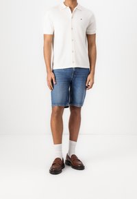 Kremowa koszulka polo z krótkim rękawem i guzikami, zestawiona z niebieskimi dżinsowymi szortami. Dopełnieniem stylizacji są brązowe mokasyny i białe skarpetki do kostki.