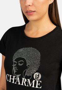 T-shirt nera con una grafica di un volto femminile con afro, impreziosita da strass, e la parola "CHARME" in testo bianco.