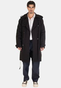 Cappotto nero con chiusura doppiopetto, ampie rever, dettagli con bottoni e tasche laterali, abbinato a jeans in denim larghi e sneakers.