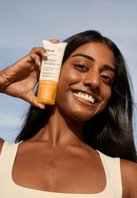 Sonnencreme-Feuchtigkeitscreme von Murad in einer Tube mit gelbem Boden und weißem Deckel, gehalten von einer Person mit langen dunklen Haaren vor blauem Himmel.