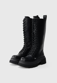 Melnas ādas ceļgala augstuma zābaki ar šņorēm priekšā, masīvu gumijas zoli un cilpu aizmugurē. Gluda tekstūra un izturīgs dizains.