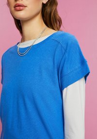 Jeune femme porte un haut bleu à manches courtes par-dessus une chemise blanche à manches longues et un collier chaîne en argent sur un fond rose.