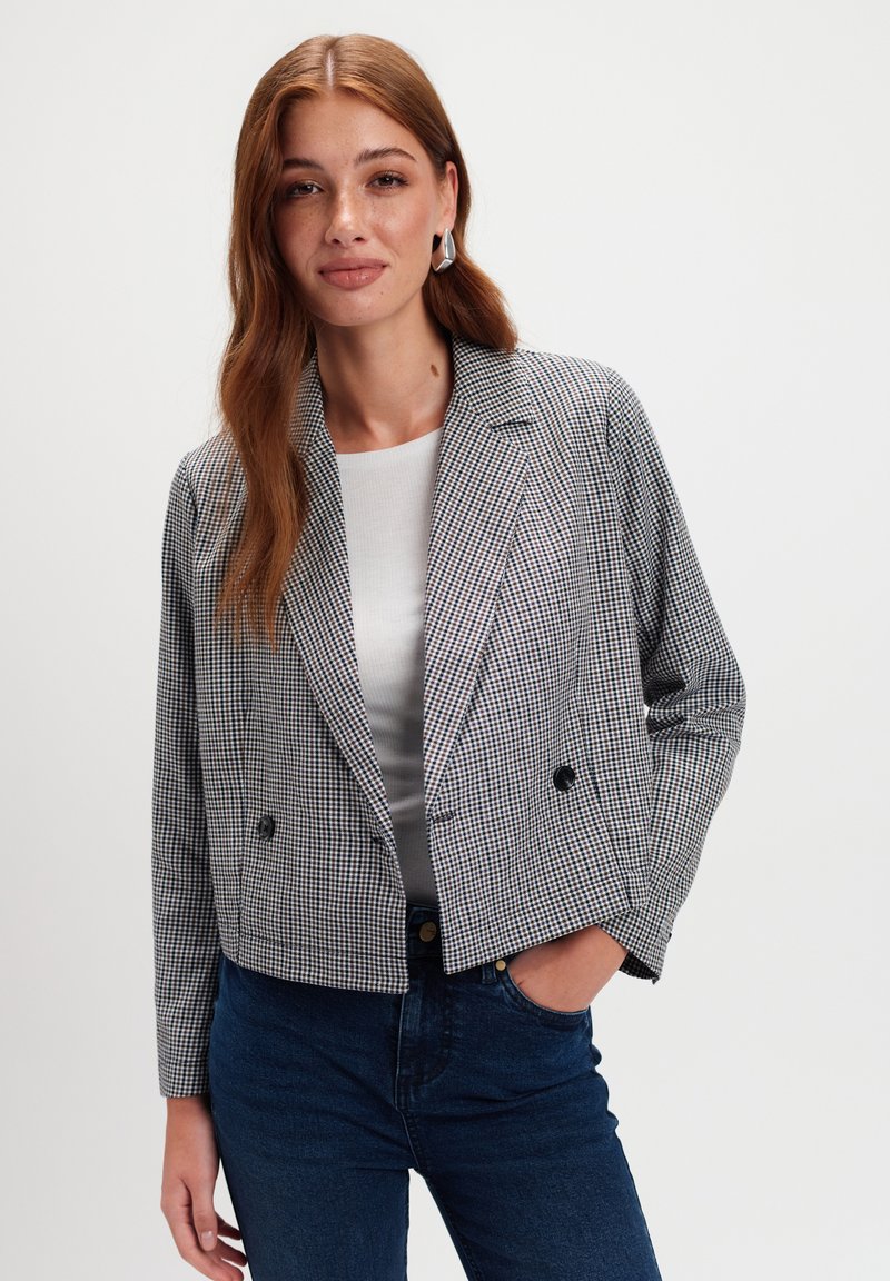 Młoda kobieta z długimi czerwonymi włosami, nosząca srebrne kolczyki, w kratkowany krótki żakiet, białą bluzkę i ciemnoniebieskie dżinsy, z ręką w kieszeni, na neutralnym tle.