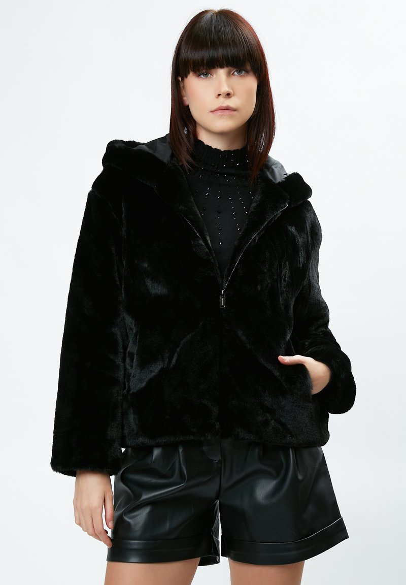 Sort faux fur jakke med hætte, lynlås lukning og brede ærmer. Parret med sort læder shorts. Glat tekstur, ingen bemærkelsesværdige mønstre.