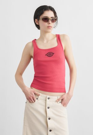 Vrouw draagt een rode Dickies tanktop, een beige rok met knopen en een zonnebril, poserend met haar handen in de zakken van de rok tegen een effen achtergrond.