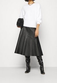 Weißer, flauschiger Pullover, schwarze Midi-Lederrock mit Nähten, kniehohe schwarze Stiefel und eine schwarze Clutch-Tasche. Detail mit klobiger Kettenkette.