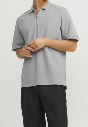 Person wearing a light grey short-sleeve polo shirt with a half-zip collar and black pants, hands gently clasped at waist.