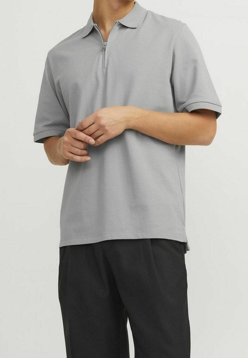 Personne portant un polo à manches courtes gris clair avec un col à demi-fermeture éclair et un pantalon noir, les mains délicatement jointes à la taille.