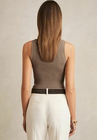 Top a costine senza maniche in taupe, abbinato a pantaloni bianchi sartoriali e una cintura nera. La texture liscia e il design aderente esaltano la silhouette.