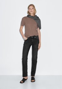 Brown short-sleeve t-shirt paired with black straight-leg jeans. Model wears a gray scarf draped over shoulders and black sandals. Simple design.
