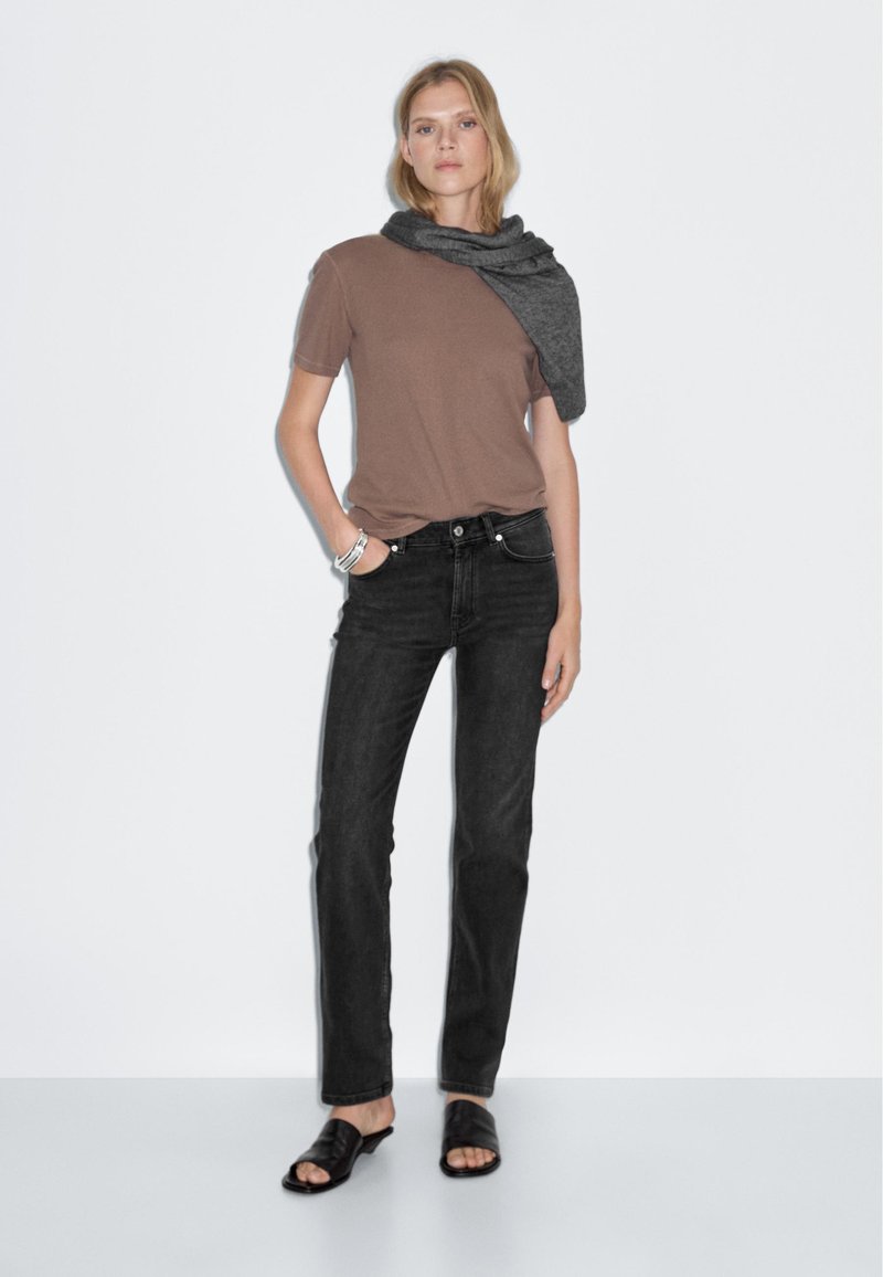 Brown short-sleeve t-shirt paired with black straight-leg jeans. Model wears a gray scarf draped over shoulders and black sandals. Simple design.
