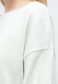 Világosszürke pulóver puha, texturált anyagból, laza, széles nyakkivágással és lelógó vállvarrással.