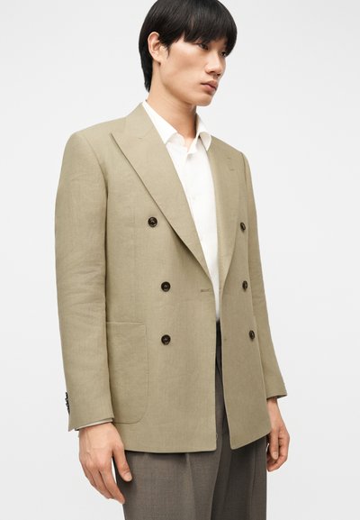 Άνδρας φοράει μπεζ σακάκι διπλού στησίματος με σκοτεινά κουμπιά, λευκό πουκάμισο και γκρι παντελόνι, στέκεται μπροστά σε απλό φόντο.