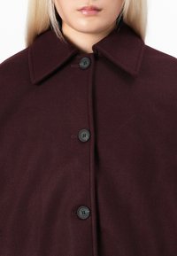 Personne à la peau claire et aux cheveux blonds portant un manteau bordeaux foncé boutonné avec trois boutons noirs et un large col.
