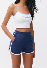 Haut court blanc côtelé avec bordure bleue et texte "MALIBU" ; associé à un short bleu marine avec des accents blancs sur les côtés et une taille élastique.
