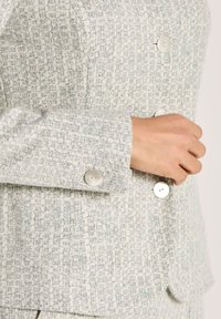 Hand, die einen Knopf an einem hellgrauen strukturierten Blazer mit großen weißen Knöpfen und subtilem Karomuster anpasst.