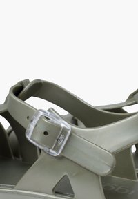 Olive green sandals with a flexible material, featuring cross straps and a clear plastic buckle for adjustment, showcasing modern design elements.