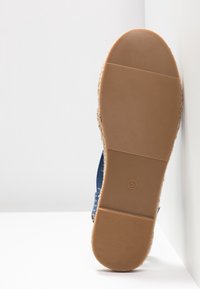 Semelle d'une chaussure en tissu bleu avec une semelle extérieure en caoutchouc beige marquée taille 5, positionnée verticalement contre un coin de mur blanc.