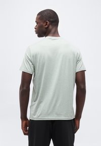 Άνδρας με πλάτη, που φοράει ανοιχτό πράσινο κοντομάνικο μπλουζάκι και μαύρο παντελόνι, μπροστά σε απλό λευκό φόντο.