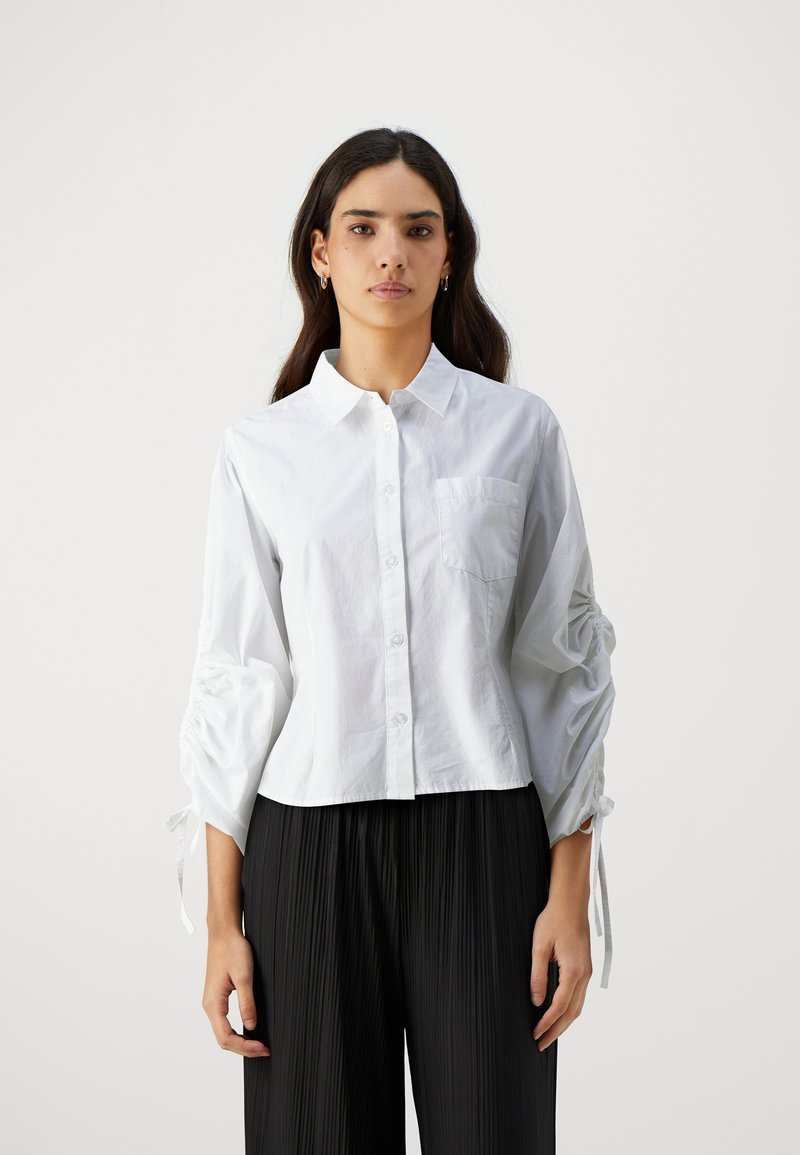 Blusa bianca con bottoni davanti, colletto e tasca sul petto. Presenta maniche arricciate con lacci. Indossata con pantaloni neri a pieghe.