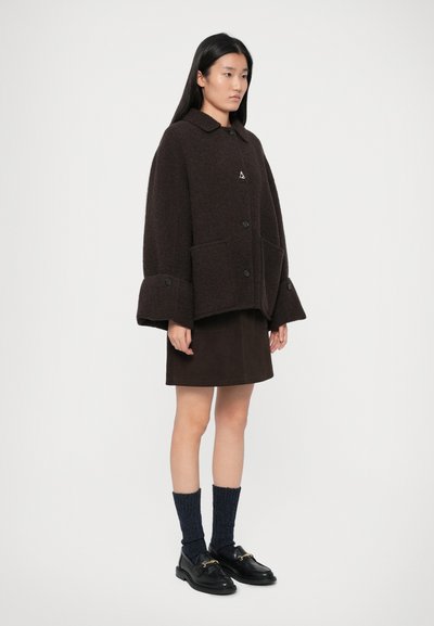 Sieviete, tērpusies tumšā oversized pogājamā mētelī, savienotajā svārku, tumši zilu zeķu un melnās loafers ar zelta detaļām, stāvot pret vienkāršu fona.
