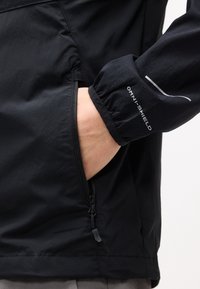 Main dans la poche latérale d'une veste noire avec une fermeture éclair visible et "OMNI-SHIELD" imprimé sur le bord élastique de la manche.
