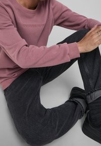 Maglione rosa chiaro con logo "JACK&JONES" in rilievo, abbinato a jeans in denim scuri e svasati. Sneakers nere in vista. Tessuto dalla superficie liscia.