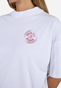 T-shirt en coton blanc avec un logo graphique rond en rouge portant le texte "DOLCE VITA PEGADOR." L'encolure est haute et côtelée, avec des manches courtes.