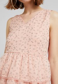 Haut rose sans manches avec un décolleté en V, présentant un ourlet péplum superposé et un motif floral. Le tissu est léger et légèrement transparent.