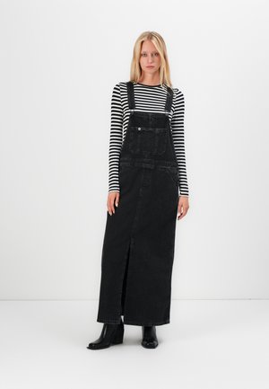 Schwarzes Denim-Maxikleid mit verstellbaren Trägern, Fronttasche und Schlitzdetails. Kombiniert mit einem schwarz-weißen gestreiften Langarmshirt.