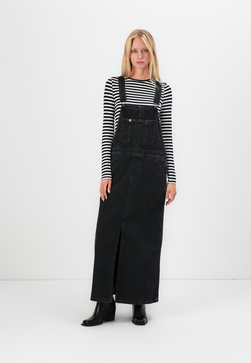 Musta denim-maksimekko, jossa on säädettävät olkaimet, etutasku ja halkioyksityiskohta. Yhdistetty musta-valkoiseen raidalliseen pitkähihasteiseen puseroon.