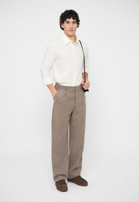 Lett hvit skjorte med oppbrettede ermer, kombinert med høytlivde, vide brune dressbukser i tweed og brune sko, holder en svart veske.