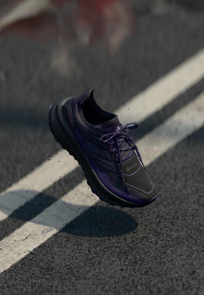 Chaussure de course violette et noire à lacets en l'air au-dessus d'une route asphaltée avec des marquages de voie blancs, projetant une ombre au sol.