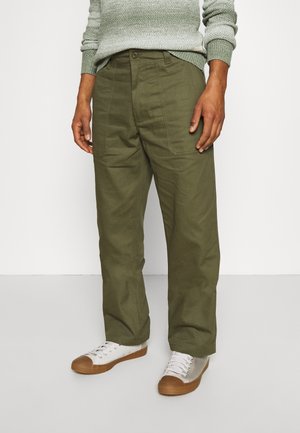 Männlich, trägt olivgrüne locker sitzende Hose mit aufgesetzten Fronttaschen, kombiniert mit weißen Low-Top-Sneakern mit braunen Sohlen.