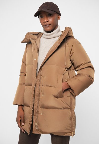 Hnedý nafukovací kabát s kapucňou, ktorý má zips, veľké predné vrecká a prešívanú textúru. Skombinovaný s béžovým rolákom.