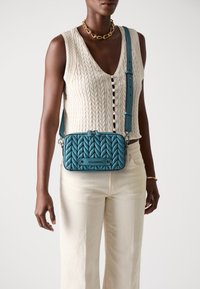 Geantă crossbody matlasată de culoare teal, cu formă rectangulară, accente din metal și curea ajustabilă, purtată peste un top crem din tricot cu model de cablu.